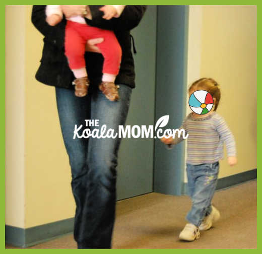 Toddler follows mom carrying a baby through a hallway for her playgroup. Photo by Bonnie Way.