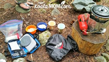 A backpacking kitchen: lightweight stove, fuel bottle, pot, utensils, and bowls with instant oatmeal and cups with instant coffee, waiting for water to boil.