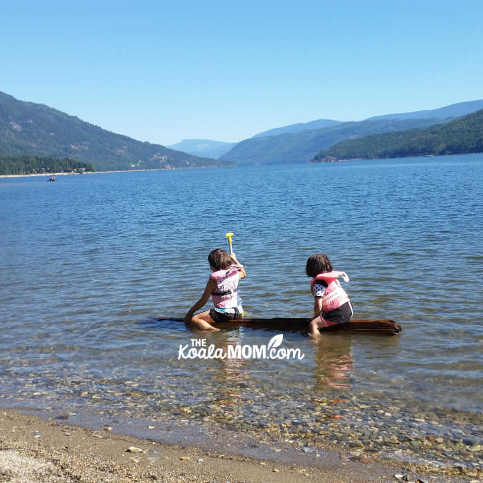 Two sisters in PFDs paddling a log on Shuswap Lake.