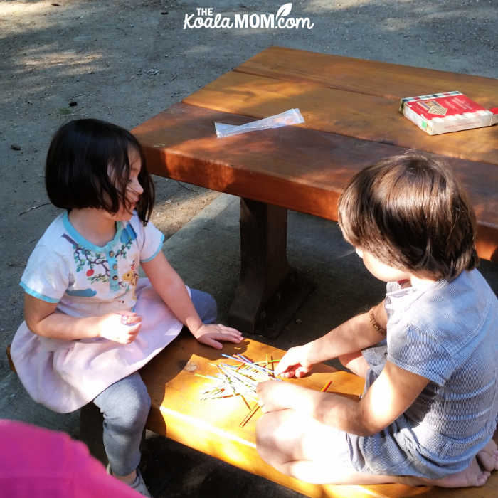 4-year-old and 7-year-old play pick-up-sticks at a picnic table.