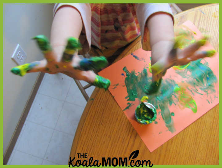 Child holding up both hands covered in finger paint above an orange piece of construction paper covered in green finger paint. Photo by Bonnie Way.